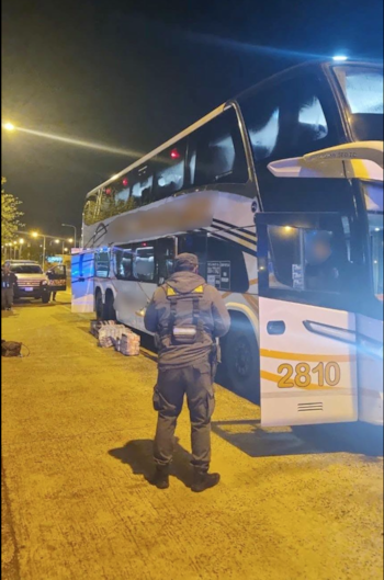 Un agente de seguridad de espaldas junto a un autobús de dos pisos y varios bultos colocados en el suelo durante la noche